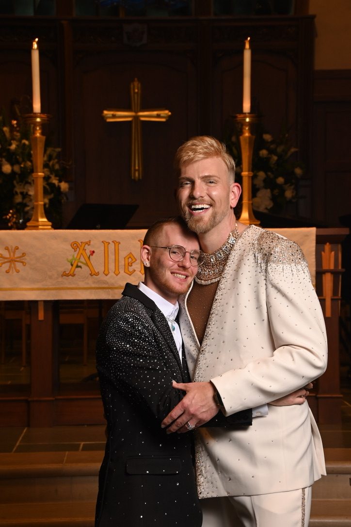 Stephen Aber in a cream embellished jacket with a statement crystal collar, posing with a friend in a church sanctuary